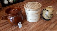 Brown pottery crackpot with vented lid, bamboo food steamer, Asian medicine cooker electric pot with Asian script and person illustration on the floor.