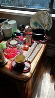 Top view of wooden table with various small decorative Asian items including fans, candle holders, coasters, hanging red braided ornaments, and beaded items arranged in natural sunlight.