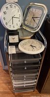 Photo showing six different clocks arranged on and around a metal and plastic storage trolley with multiple drawers.