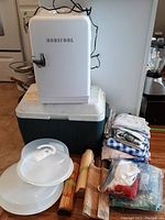 Photo showing the Mobicool white mini fridge on top of the Coleman cooler, surrounded by folded tablecloths, rolled placemats, and two covered dessert carriers.