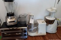 Four kitchen appliances on a countertop: Oster blender, compact black and silver toaster oven, Black & Decker FP1200 food processor, Huron HU100 juice extractor.