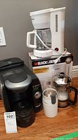 Photo showing the full lot: Bosch Tassimo coffee maker, Black & Decker white drip coffee maker on its box, white electric coffee grinder, and manual glass coffee press with metal lid and black handle, placed on a wooden floor.