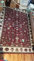 Full view of the red floor rug with floral and vine design, surrounded by a cream floral border, laid on hardwood floor with furniture and boxes nearby.