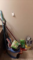 Full view of cleaning supplies lot showing floor sweepers, blue bucket with wheels, dustpans, cleaning cloth refills and sponges on the floor.