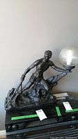 Front and right side view of a ceramic statue lamp showing a man holding a ribbed glass globe lamp. The statue is dark-colored and has scratches. The lamp globe has visible damage at the back.
