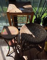 Three wicker items including square stool and two stands shown on wood floor by window with sunlight.