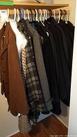 Wide view of 9 coats, jackets, vests hanging in a closet on hangers, mostly brown, black, plaid, and grey.