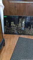 Framed artwork of New York City skyline at night with illuminated buildings, leaning against a wooden panel wall on wood floor with textured dark rug.