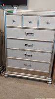 Front view of the silver chest showing five drawers: 3 small top drawers with square metal pulls, 2 larger drawers with curved handles, and a decorative gold panel on bottom drawer.