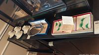 Four white Tiffany tea cups and saucers on a black surface with silver plated plate beside salt and pepper shakers; three cloisonne plates in box with protective padding.