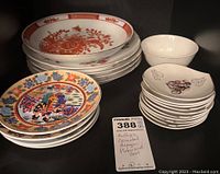 Stacked oriental plates and bowls including a set of white plates with red-orange floral designs, multicolored plates with intricate patterns, and white bowls with purple floral motifs, shown with a MaxSold tag describing the lot as multiple oriental designed plates and bowls.