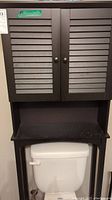Dark brown over-the-toilet storage shelf with louvered doors above open shelving, shown mounted over a white toilet. Visible dust and dirt indicating need for cleaning.