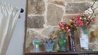 Four glass vases including two with faux pink flowers, arranged in front of a stone wall.
