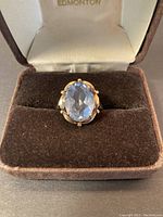 Front view of ring in jewelry box showing oval faceted blue stone and prong setting