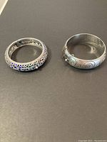 Two sterling silver bracelets on a dark background, showing both ornate enamel filigree bangle and plain scroll bangle