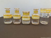 Full set of five glass containers and a glass showing hand painted yellow bands with white floral designs and gold rims.