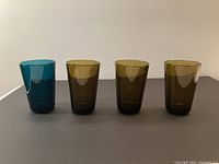 Four colored Nuutajärvi highball glasses shown from front on dark surface: one blue and three olive green glasses in a row.