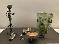 Wide view showing all items: green glass mask lamp, female bronze-style statue, candle holder, and metal figures on black background.