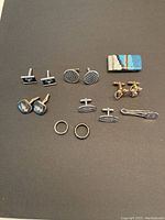 Full view of lot items: five pairs of cufflinks, two rings, one money clip and one tie clip on black surface.