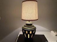 Large ceramic lamp with beige round fabric lampshade and glowing light through ceramic base cutouts.