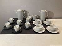 Two sets of white porcelain coffee and tea sets with black and white saucers, including tall coffee pots, cups, saucers, and creamers. One creamer has a chip.