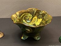 Close-up of a green iridescent Zsolnay art nouveau dish with a ruffled edge and swirled design, standing on three short feet on a dark surface.