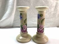 Front view of the pair of cream-colored ceramic candlestick holders with detailed hand painted purple and pink flowers, leaves around the cylindrical bodies.