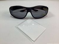Front view of black framed XL polarized sunglasses with dark lenses, displayed with folded white cleaning cloth on white surface.