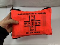 Closed orange first aid kit pouch held in hand showing first aid cross and text
