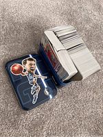 A large stack of loose Pokémon trading cards inside a blue metal tin with a caricature of a basketball player on the lid.