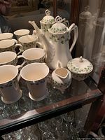 Photo showing the full tea set with teapot, cream and sugar dishes, and multiple cups in white ceramic with green shamrock design, displayed in cabinet with mirror.