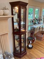 View of the tall curio cabinet placed near a fireplace and window, showing dark wood frame and glass doors with visible interior shelves and light on top.