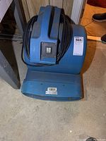 Front view showing heavy blue plastic floor blower fan with integrated top handle and power cord wrapped around it.