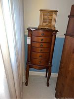 Tall wooden jewelry armoir with six drawers, round knobs, and decorative columns, standing in corner of room