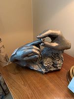 Side view of sculpture showing two hands, one holding a ring, positioned on wooden surface.