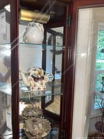 Wooden display case with a white art bowl with leaf extensions on the top shelf, floral chair-shaped tea pots below, and nest display with eggs on the bottom shelf.
