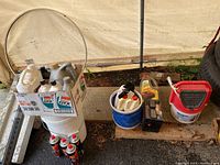 Overview photo showing a white milk crate holding various bottles of motor oil and lubricants, several butane cartridge cans on the ground, a bait bucket, and a small black battery on a wooden shelf.