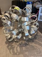 Front view of chandelier showing silver metal ribbons arranged in loops forming a spherical, modern artistic design on carpeted floor.