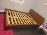Assembled wooden double bed frame with slatted mattress support, shown in room on dark wood floor with red carpet.