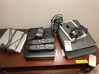Top view of Xbox console with two remote controls and various power cords on a table with two laptops and a tablet.