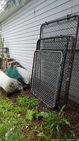 Two metal chain link gates resting against house siding outdoors with grass and soil below, showing metal frame and wire mesh.