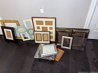 A group of over 15 assorted picture frames of varying sizes and colors displayed on the floor against a white wall.