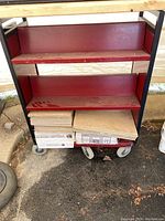 Front view of metal library cart showing three red shelves with black frame, bottom shelf holds wood pieces (not included).