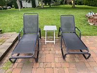 Two black patio lounge chairs with mesh fabric and metal frames positioned on brick patio with a small white folding side table between them. Outdoor backyard setting visible in background.