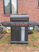 Front view of Master Chef gas BBQ grill showing five orange control knobs, side shelves, storage cabinet, and overall structure on outdoor brick surface.