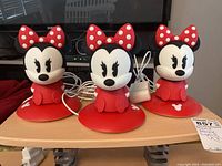 Three identical Minnie Mouse nightlights on a wooden surface in front of a TV screen, with power cords attached and bundled at the back.