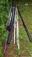 Various assorted metal and plastic pipe pieces of different lengths and colors (black, white, gray) laid on the grass outdoors near bushes.