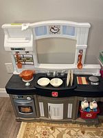 Front view of white, black, and gray plastic child's play kitchen with included dishes and utensils on countertop and hanging hooks.