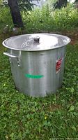 Side view of a large aluminum pot with riveted handles and a lid with a black knob, placed outdoors on grass.