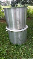 Two stacked large aluminum pots placed on grass outdoors, showing outer surface and handles.